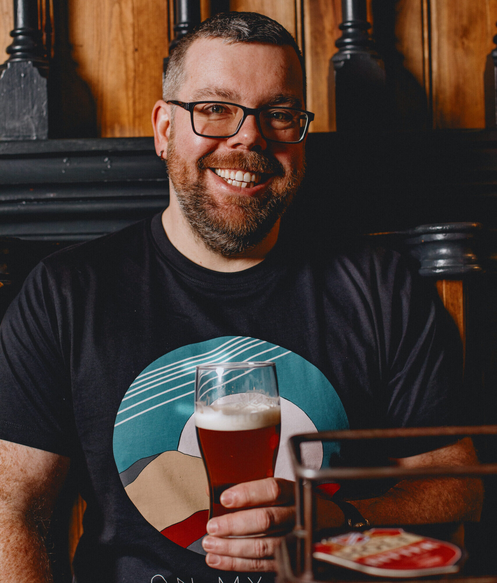 A man smiling sat down at a table holding a pint.