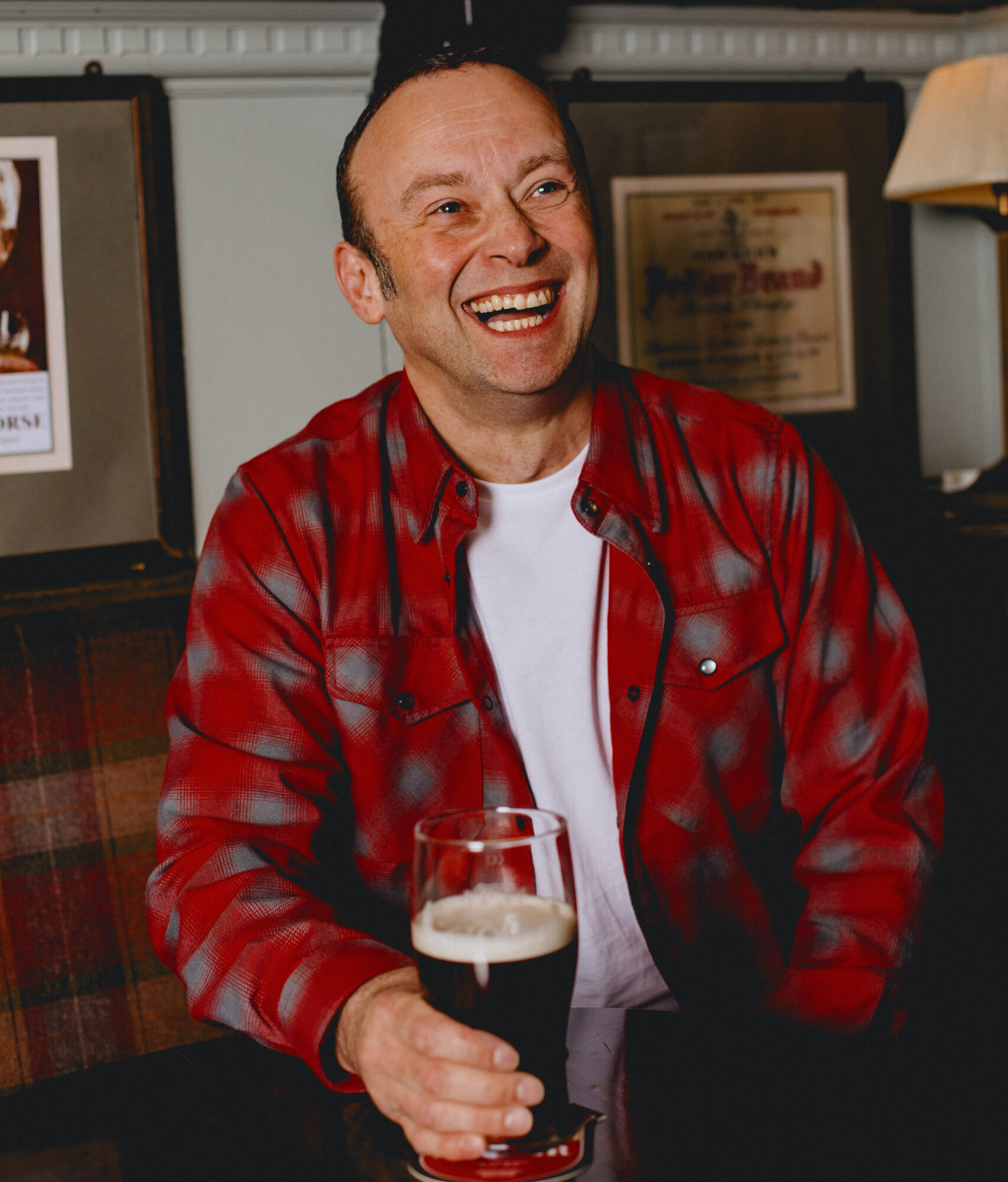 A man laughing sat down holding a pint.